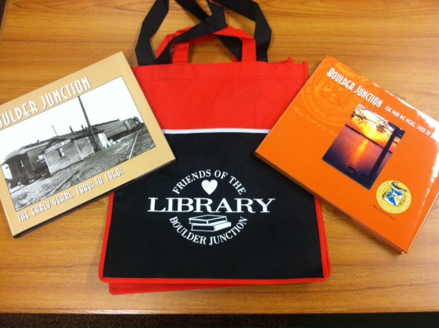 A red and black library tote bag flanked by two books titled "Boulder Junction" on a wooden table.