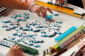 A game of Mahjong in progress with hands reaching for tiles on a table surrounded by colorful racks.