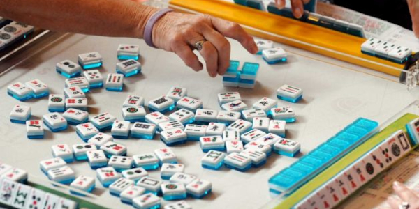 A game of Mahjong in progress with hands reaching for tiles on a table surrounded by colorful racks.